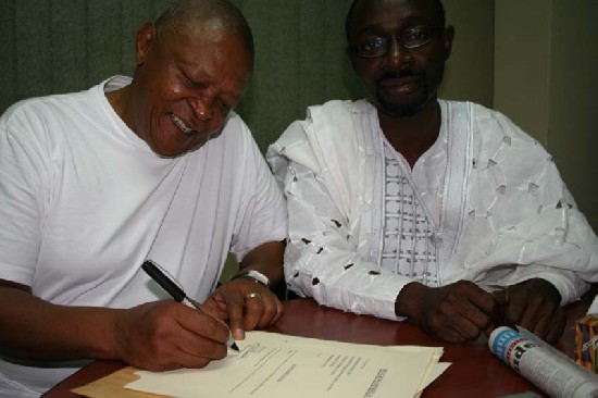 happily signing documents to confirm his membership of the Board of the Woyome Foundation for Africa. Looking on is te Board Chairman and Ag. Project Manager Alfred Woyome